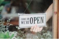Ladeninhaberin heißt Kunden mit einem Schild auf Englisch willkommen: Come in! We're open